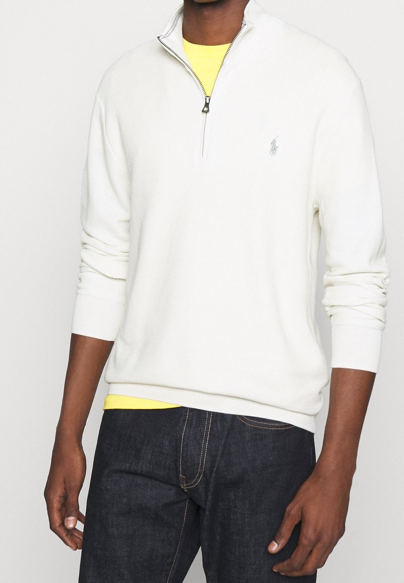 Weißer Strickpullover mit einem Viertel-Reißverschluss-Kragen, gerippten Bündchen und Saum, über einem gelben Hemd getragen. Dunkelblaue Denim-Jeans sind sichtbar.