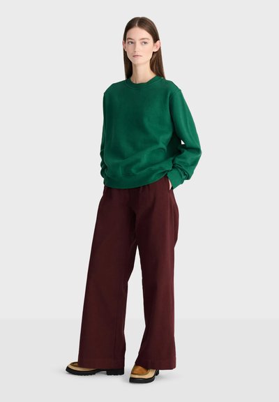 Femme debout portant un sweat-shirt vert, un pantalon large rouge foncé et des chaussures beige avec des semelles noires sur un fond clair uni.