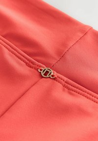 Nærbillede af koralfarvet stof med detaljeret syning og lille rund metalspænde med geometrisk design.