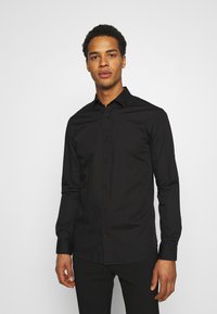 Camisa negra de manga larga, ajustada, con cuello abotonado y botones en la parte delantera. Textura suave con un diseño liso, combinada con pantalones negros.