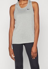 Kobieta w beztaliowym szarym topie Nike na ramiączkach i czarnych legginsach, stojąca z ramionami wzdłuż ciała na jednolitym białym tle.