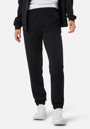 Osoba ubrana w czarne spodnie dresowe z elastycznymi mankietami, białe trampki, czarną kurtkę zapinaną na zamek i białą koszulę na tle jednolitego tła.