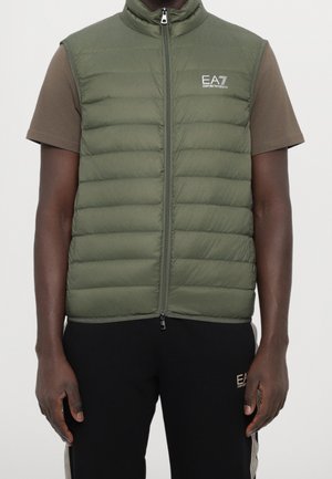 Man die een olijfgroene donsjack met EA7-logo draagt, een bruin shirt met korte mouwen en zwarte EA7-trainingsbroek met strepen aan de zijkant.