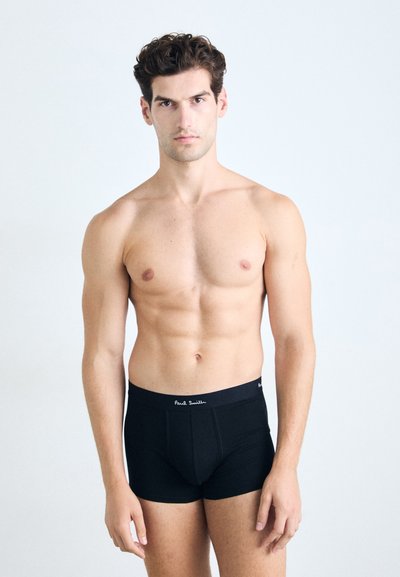 Boxer shorts en coton noir à coupe près du corps, avec une ceinture de marque. Un modèle avec des cheveux courts et ondulés se tient devant un fond clair.