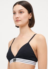 Black bralette made of soft fabric with a triangular shape, thin straps, and a white striped band featuring a logo at the center front.