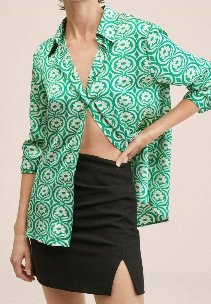 Blouse verte à motifs floraux, col ouvert et manches longues, associée à une mini-jupe noire avec une fente latérale.