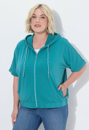 Lyshåret kvinde iført teal lynlåshoodie med hvide snore og blå jeans, stående med den ene hånd på hoften mod ensfarvet baggrund.