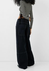 Vaqueros de pierna ancha en denim oscuro con costuras prominentes, con bolsillos traseros y un acabado texturizado. Combinados con una blusa de rayas en blanco y negro.