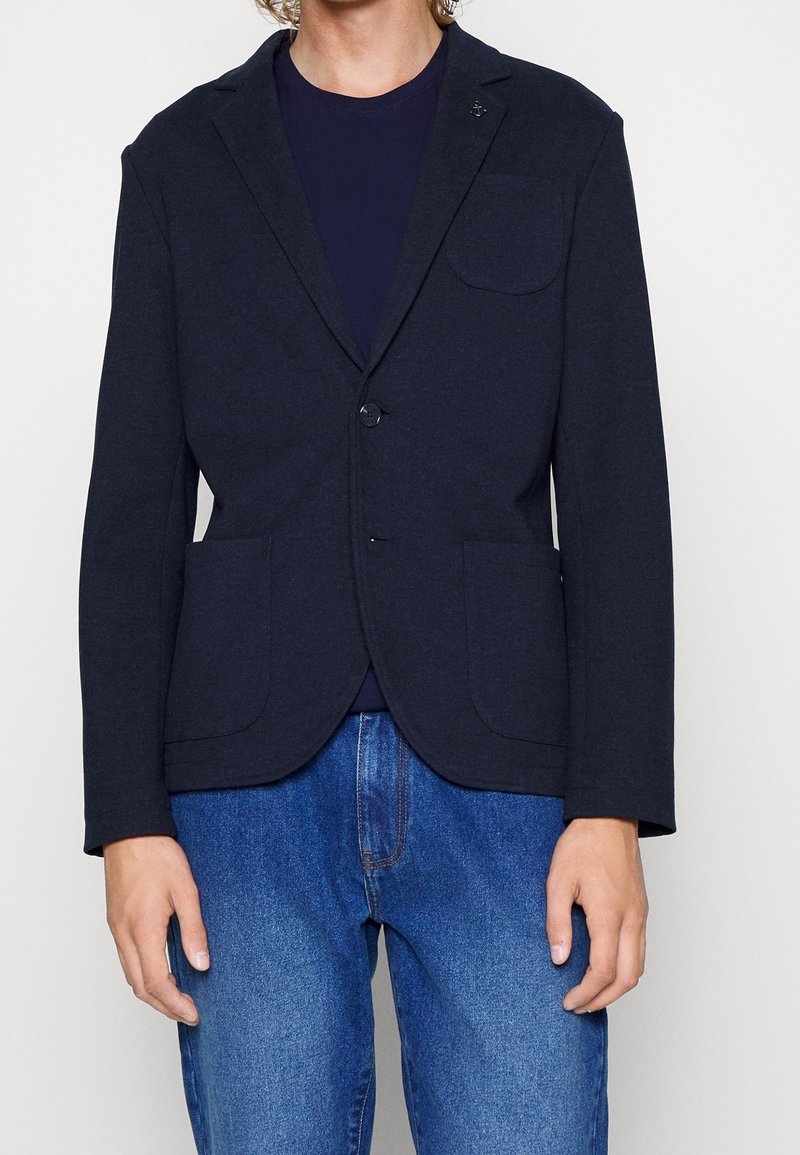 Personne portant un blazer bleu marine foncé par-dessus une chemise bleu marine et un jean bleu, avec le blazer boutonné au milieu et les mains détendues le long du corps.