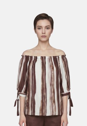 Vrouw met kort haar die een off-shoulder blouse draagt met verticale bruin-witte strepen en gebonden mouwen, staand tegen een effen achtergrond.