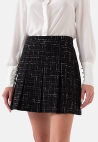 Zwarte plissé rok van een gestructureerde stof, met witte ruiten. Gecombineerd met een witte blouse met knopenmanchetten.