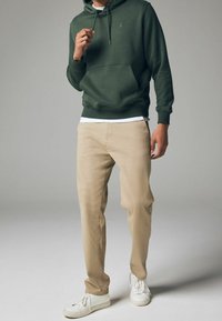 Homme portant un sweat à capuche vert foncé, un pantalon beige et des baskets blanches, debout devant un fond gris uni.