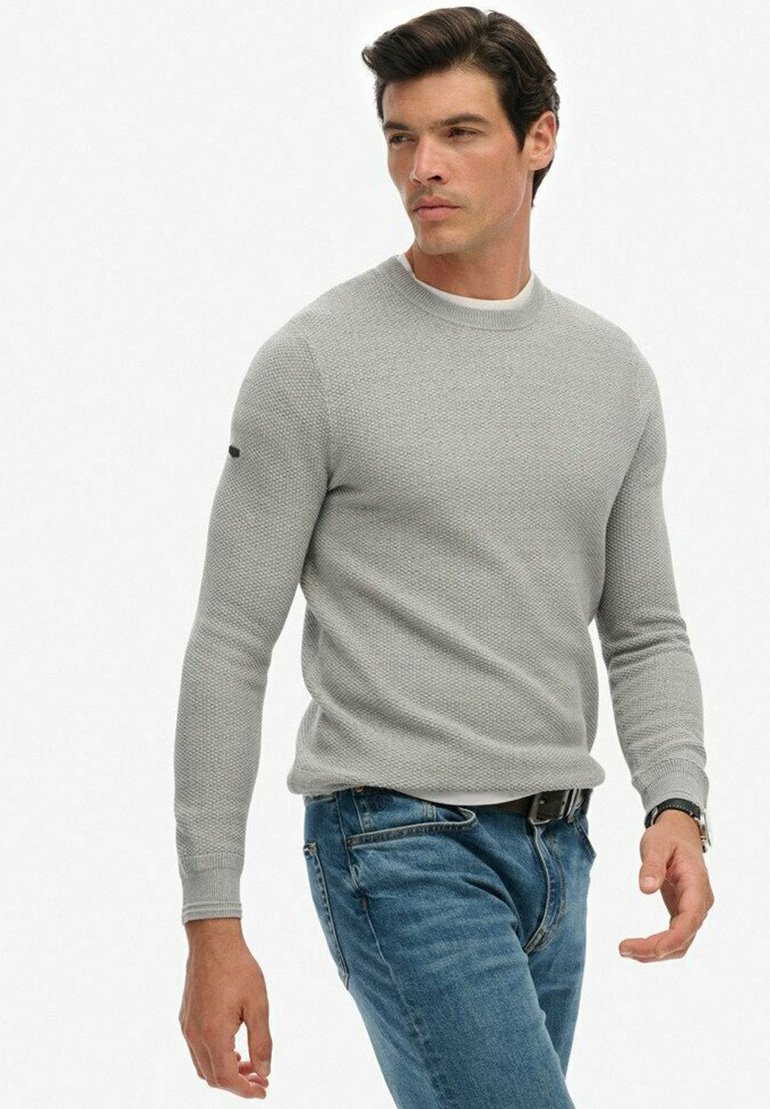 Mężczyzna w jasnoszarej, teksturowanej swetrze i niebieskich dżinsach, idący z skoncentrowanym wyrazem twarzy na tle białego tła.