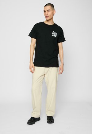 T-shirt in cotone nero con una grafica di una rosa bianca sul petto sinistro. Indossato con pantaloni color crema e scarpe nere, caratterizzato da una vestibilità casual.