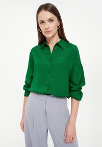 Grüne knopfnahe Bluse mit einer weichen, glatten Textur und hochgekrempelten Ärmeln, kombiniert mit hellgrauen, gefalteten Hosen. Minimale Hardware-Details.
