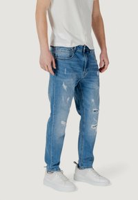 Jeans de mezclilla azul claro con un ajuste relajado, que presentan detalles desgastados y rotos. Combinados con zapatillas blancas y una camisa de mangas cortas.