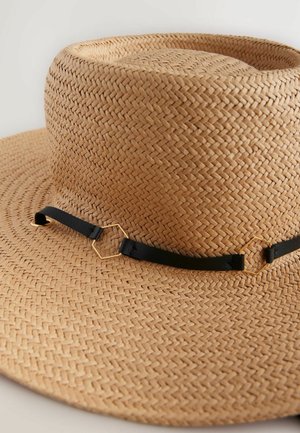 Sombrero de paja trenzada con ala ancha, que presenta una banda de cuero negro y un acento hexagonal dorado. Textura natural en color beige claro.
