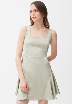 Kobieta z długimi brązowymi włosami w bezrękawowej sukience w kształcie litery A z satyny w jasnym kolorze szałwii, stojąca na tle jednolitego białego tła.