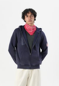 Sudadera con cremallera color navy, con bolsillos frontales, fabricada en algodón y con puños acanalados. Presenta un pequeño parche de la bandera estadounidense en la manga izquierda.