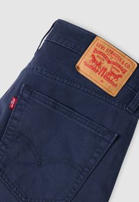 Τζιν παντελόνια σε σκούρο μπλε χρώμα με διακριτικό σχέδιο στην πίσω τσέπη, που διαθέτει καφέ δερμάτινη ετικέτα με λεπτομέρειες της μάρκας και κόκκινη ραφή ως διακοσμητικό στοιχείο.