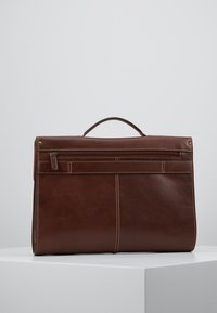 Serviette en cuir marron avec poignée supérieure, poche avant zippée et détail de couture verticale, placée sur une surface blanche avec un fond gris.