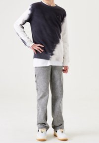 Dunkelgrauer und weißer Batik-Pullover mit gerippten Bündchen, über einem weißen Lagen-Shirt getragen, kombiniert mit hellgrauen geraden Hosen und Sneakers.