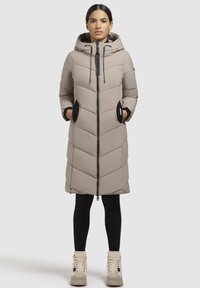 Langer, hellbeige Daunenmantel mit Chevron-Muster, verstellbarer Kapuze, Reißverschluss und schwarzen Akzenten an den Taschen. Getragen über schwarzen Leggings.