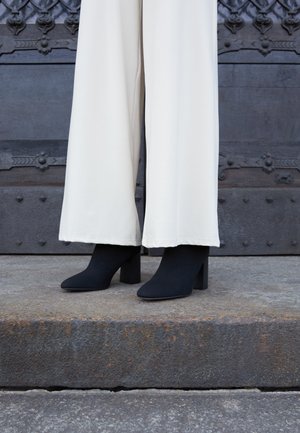 Black heeled ankle boots worn with white wide-leg pants standing on stone steps in front of a dark ornate metal door.