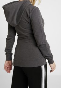 Szara bluza z kapturem wykonana z miękkiego materiału, z długimi rękawami, ściągaczami na mankietach i kieszenią kangurka, noszona z czarnymi spodniami.