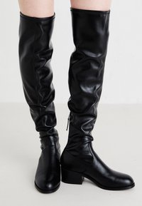 Botas negras sobre la rodilla, elaboradas en cuero suave, con un diseño holgado, tacón bajo y detalle de cremallera lateral para facilitar su uso.