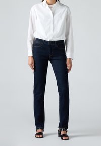 Personne portant une chemise blanche boutonnée, un jean slim bleu foncé et des sandales noires à talons ouverts, debout devant un fond uni.
