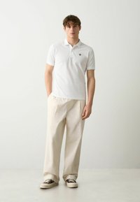 Cămașă polo albă cu mâneci scurte, cu logo brodat pe piept, asortată cu pantaloni bej deschis, plisati și adidași alb-crem.