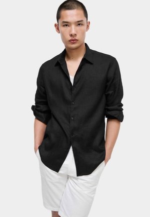 Junger Mann mit kurzem Haar, der ein schwarzes Hemd mit aufgerollten Ärmeln und weiße Shorts trägt, stehend mit den Händen in den Taschen.