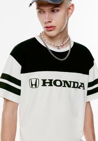 Valkoinen t-paita mustilla raidoilla ja "HONDA"-logolla paksulla mustalla fontilla. Sisältää lyhyet hihat ja rento istuvuus.