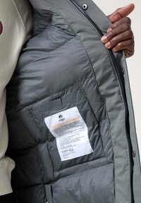 Chaqueta acolchada gris con cierre delantero, que muestra un interior suave, una etiqueta con detalles sobre el cuidado de la tela y un bolsillo interior.