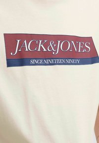 Balta medvilninė marškinėliai su stačiakampiu maroon ir jūros mėlynumo grafiniu dizainu. Tekstas skaito "JACK & JONES" ir "Nuo 1990-ųjų".