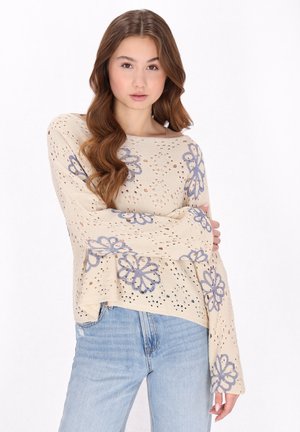 Beige Strickpullover mit großer blauer Blumenstickerei, offenes Gewebedesign und langen Glockenärmeln, kombiniert mit hellblauen Jeans.