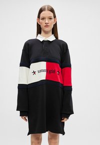 Čierne oversized polo šaty s bielymi, červenými a námorníckymi horizontálnymi pruhmi. Obsahujú vyšívané logo "tommy girl" a klasický golier.