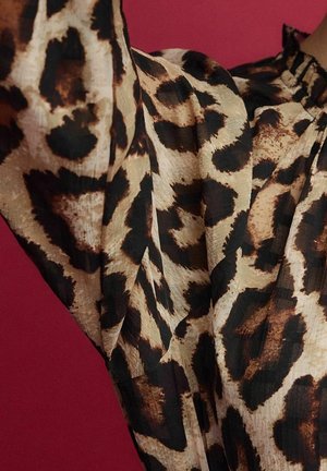 Blusa de estampado de leopardo con un tejido semitransparente y texturizado que presenta patrones en negro y marrón sobre un fondo claro beige. Detalle de escote con volantes.
