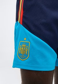 Μπλε και ναυτικό σορτς αθλητικά με κόκκινες και κίτρινες ρίγες, με κίτρινη εθνόσημο της ποδοσφαιρικής ομάδας της Ισπανίας κοντά στο τελείωμα.