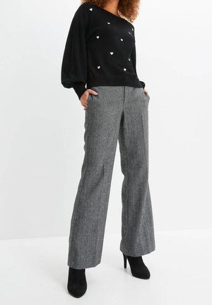 Femme portant un pull noir avec des motifs de cœurs blancs, un pantalon gris à jambes larges et des bottines noires à talons, les mains dans les poches.