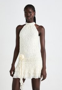 Donna con capelli a treccia che indossa un abito bianco in pizzo con scollatura a halter e dettagli floreali in vita, in piedi contro uno sfondo chiaro e semplice.