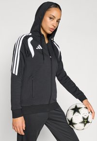 Μαύρο φούτερ με φερμουάρ, λευκές ρίγες, κουκούλα με κορδόνι και δύο μπροστινές τσέπες. Το μοντέλο κρατά μια ασπρόμαυρη μπάλα ποδοσφαίρου με σχέδιο.