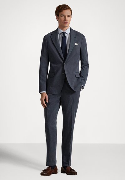 Vyras vilki tamsiai pilką kostiumą, šviesiai languotą marškinius, tamsiai mėlyną kaklaraištį, baltą kišeninę skiautę, rudas batelius su kutais ir nešioja laikrodį ant riešo, stovėdamas prieš baltą foną.