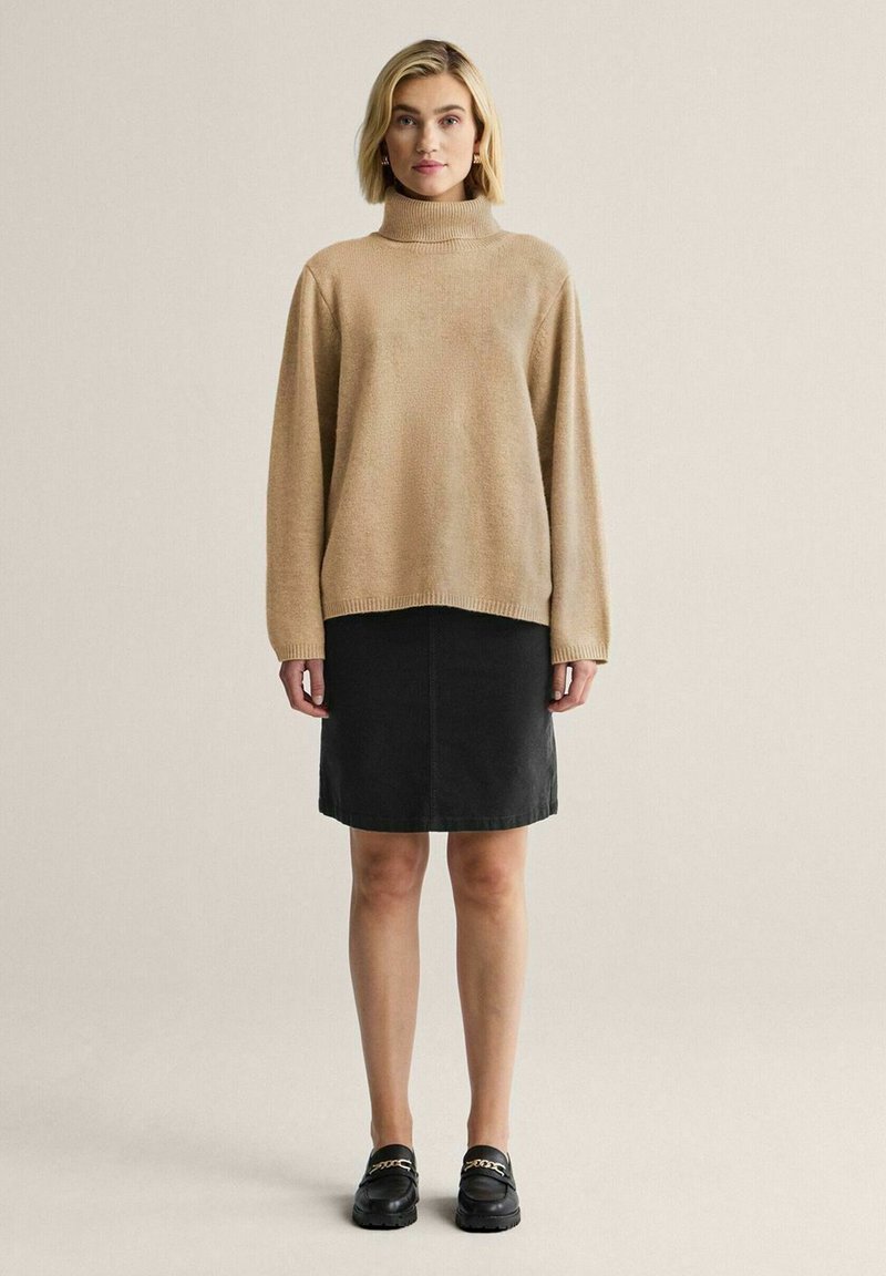 Beige stickad tröja med hög krage, kombinerad med en svart jeanskjol. Modellen bär svarta loafers med kedjedetalj. Mjuka texturer.