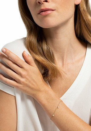 Goldene Kettenarmband mit drei kleinen runden Anhängern, am Handgelenk vor einem hellen Stoffhintergrund getragen. Glatte Textur, zartes Design.