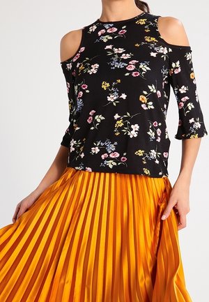 Kvinde iført en sort kold-skulder blomstret top kombineret med en lys orange plisseret nederdel, der holder nederdelen med den ene hånd.