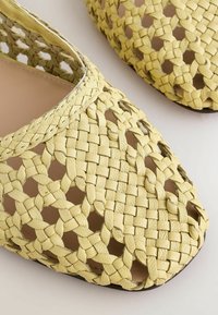 Chaussures à enfiler en cuir tissé jaune avec un motif en treillis ouvert et des orteils carrés arrondis sur un fond clair.