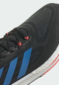 Chaussure de sport noire avec logo à trois bandes bleues, partie supérieure texturée, lacets rouges et semelle blanche avec motif de crampons.