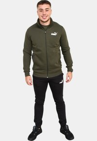 Giacca a zip verde oliva con colletto alto, abbinata a pantaloni neri aderenti. Entrambi presentano accenti con il logo bianco Puma. Il modello indossa sneakers nere.